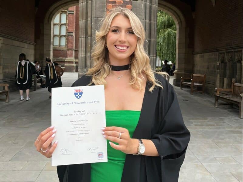 A photograph of a blonde woman holding a university certificate on graduation day.